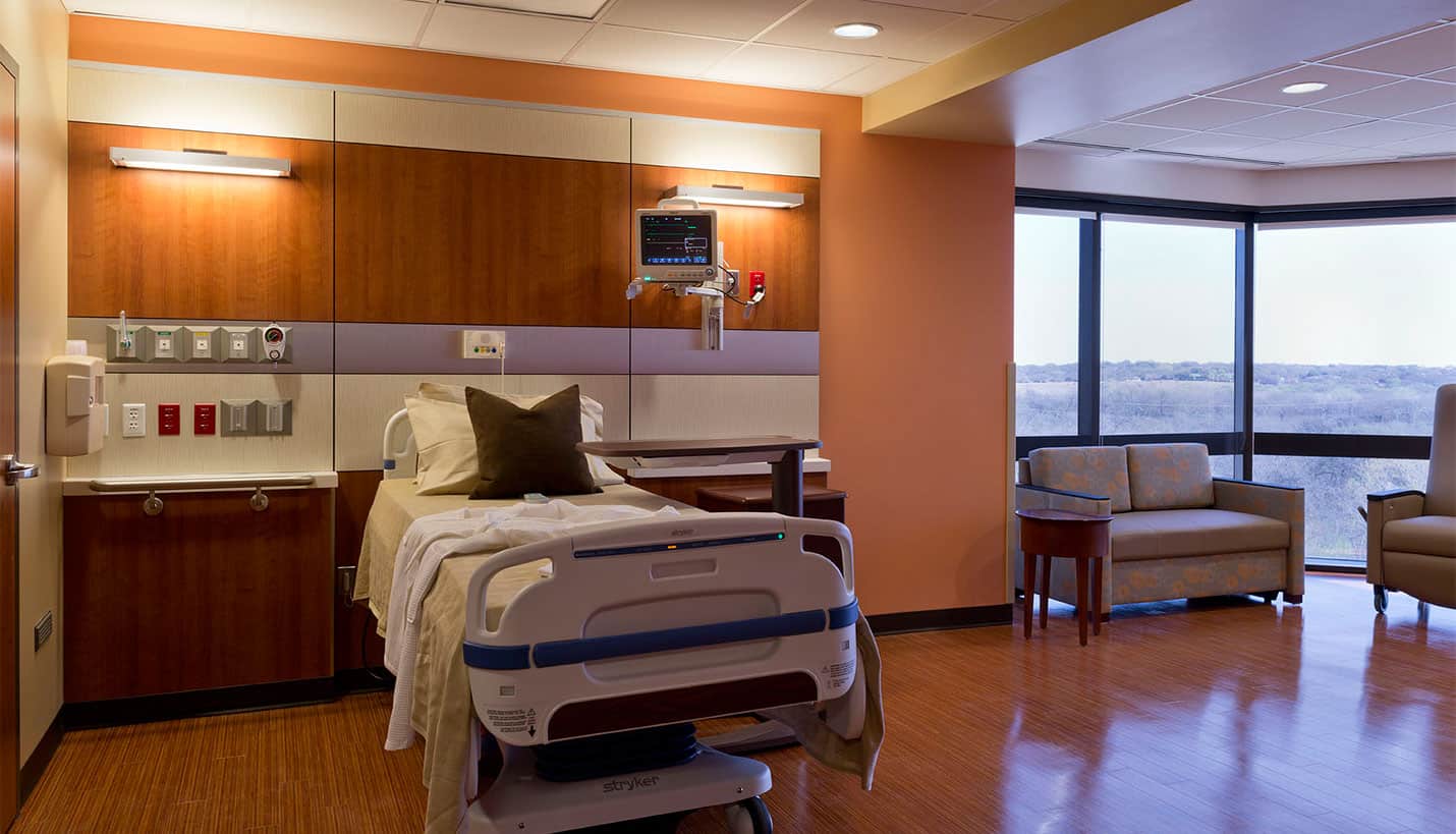 An empty hospital patient room with a bed, a window, and two couches.